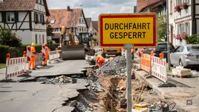 Straßenbauarbeiten mit gelbem Durchfahrt-gesperrt-Schild, Bauarbeitern in Warnkleidung und Baumaschinen in einer Ortschaft