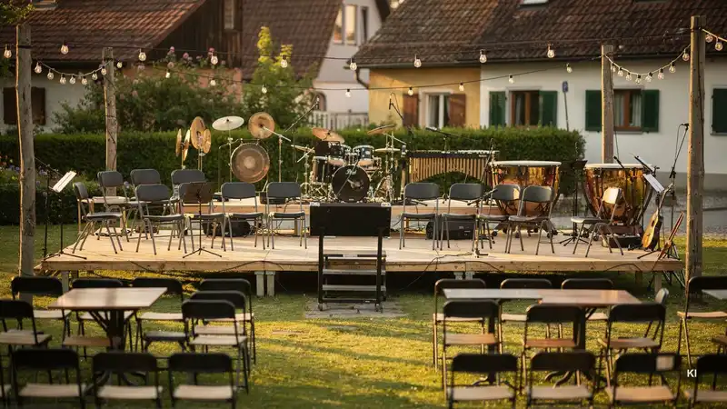 Freilichtbühne mit Schlagzeug, Percussion-Instrumenten und Stühlen, umgeben von Lichterketten und Zuschauerplätzen im Garten.